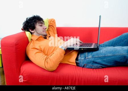 Giovane uomo vestito casualmente la posa rilassata su un divano rosso con un laptop Foto Stock