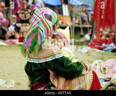 Vista posteriore del fiore seduto donna Hmong in abito tradizionale con un bambino sul suo grembo, presso il mercato del sabato, può cau, NW Viet Nam Foto Stock