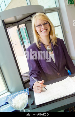 Sorridente giovane donna caucasica con lunghi capelli biondi, in un'elegante giacca blu, firmando al banco della reception di un moderno edificio di uffici. Foto Stock