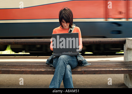 Giovane donna seduta su una panchina in una stazione ferroviaria piattaforma digitando su un computer portatile come un treno passa Foto Stock