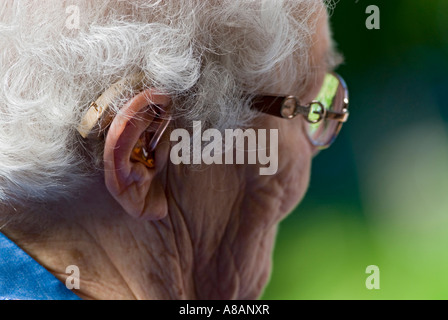 Donna anziana che indossa apparecchi acustici e occhiali all'aperto in un luminoso giardino soleggiato Foto Stock