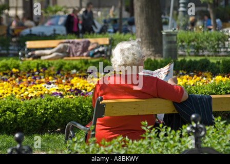 Budapest, Ungheria. Vecchia donna lettura rivista in piazza nel centro citta'. L'uomo addormentato sul banco di lavoro in background Foto Stock