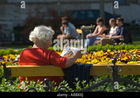 Budapest, Ungheria. Vecchia donna lettura rivista in piazza nel centro citta'. I giovani sul banco di lavoro in background Foto Stock