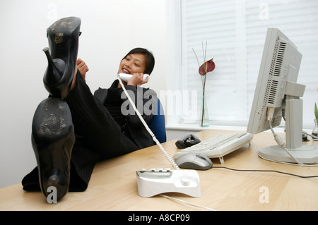Donna orientale sul telefono con piedi sulla scrivania rilassante nella parte anteriore del monitor del computer in ufficio Foto Stock