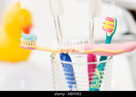 Quattro spazzolini da denti colorati in un vetro che uno di loro con il dentifricio attraverso il vetro con gomma gialla anatra in background Foto Stock