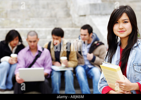 Ritratto di una giovane donna in possesso di un libro con i suoi amici seduti in background Foto Stock