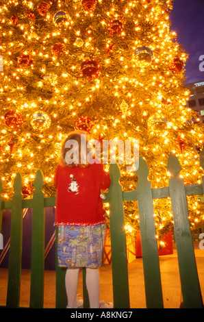 Bambina con un maglione rosso in piedi su un verde Picket Fence staring fino a un enorme vivacemente albero di Natale illuminato in Union Square di San Francisco. Foto Stock