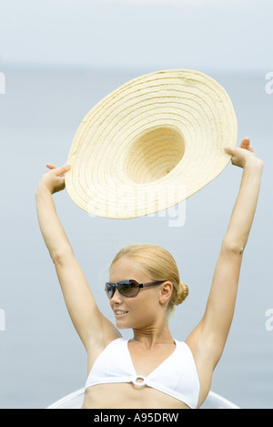 Giovane donna che indossa gli occhiali da sole, tenendo alto cappello, testa e spalle Foto Stock