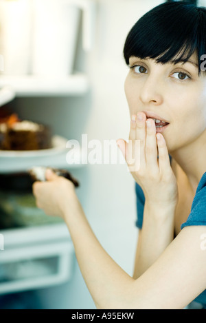 Giovane donna prendendo fetta di torta dal frigorifero, guardando la telecamera, bocca di copertura Foto Stock