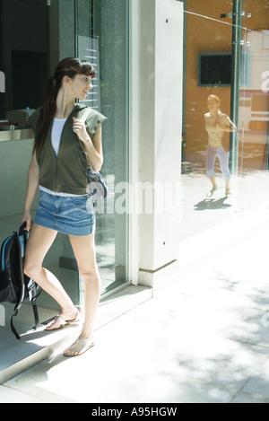Giovane donna in piedi da ingresso, guardando sopra la spalla a amico arrivando, riflessa nella finestra Foto Stock