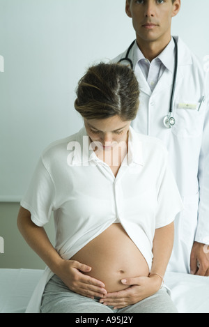 Donna incinta seduta sul lettino, guardando in giù a stomaco, dottore in piedi dietro di lei Foto Stock