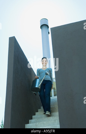 Giovane donna a piedi giù per le scale, portando annaffiatoio Foto Stock