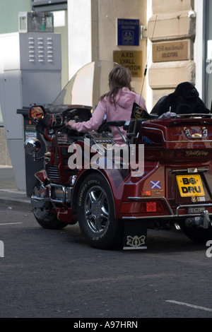 Donna con capelli biondi seduto su una grande ruota tre moto parcheggiata sul lato della strada a Dundee, Regno Unito Foto Stock