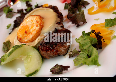 Bistecca alla griglia con formaggio fuso e uova fritte, guarnita con verdure miste, fette di cetriolo e peperone su un piatto bianco. Foto Stock
