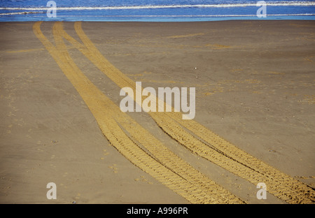 Un set di pneumatici o cingoli attraversando una chiara spiaggia sabbiosa e scomparire in o emergente da un delicato blu del mare Foto Stock