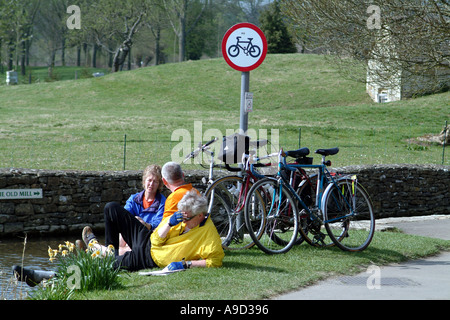 I ciclisti si prendono una pausa sulla sponda del fiume a Lower Slaughter Gloucestershire in Cotswolds England Regno Unito Regno Unito Foto Stock