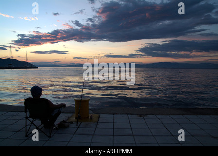 Uomo seduto nella sedia sulla banchina del porto di pesca con un asta di fronte al mare e terraferma distanti. Crepuscolo. Thassos Grecia 2006 Foto Stock