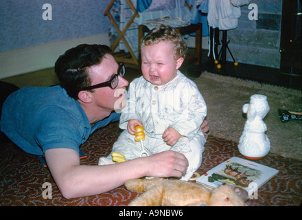 Il giovane padre caucasico si trovava sul pavimento accanto a un bambino che piangeva seduto sul pavimento in una tipica casa degli anni '1960. REGNO UNITO. Archivia immagine Foto Stock