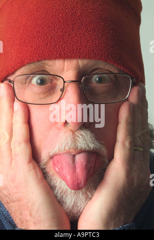 Uomo anziano in warm Red hat con le mani per affrontare dover praticare la lingua di fuori Foto Stock