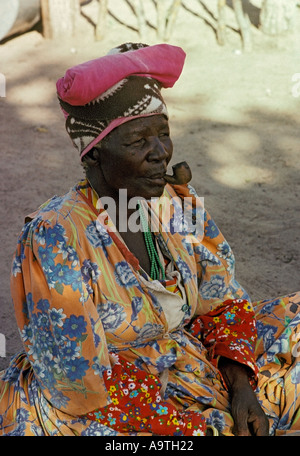 Herero donna che indossa gli abiti tradizionali e copricapo di fumare un tubo Foto Stock
