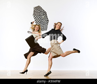 Due donne che saltava e saltando in conservativa Ufficio Wear uno portando un breve caso gli altri con un ombrello Foto Stock