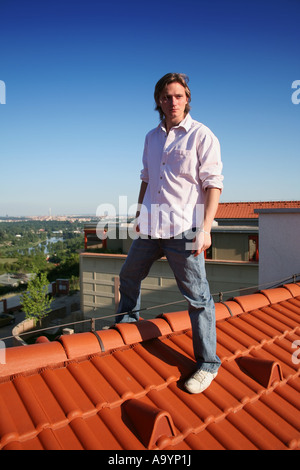 Giovane uomo sicuro di sé in piedi su un tetto di tegole rosse, che si affaccia su un paesaggio urbano panoramico sotto un cielo blu brillante. Foto Stock