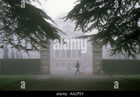 Chiswick House nel gennaio nebbia incorniciato da alberi di cedro, uomo a camminare in gateway marrone con cane al guinzaglio e in bianco e nero alle spalle del cane, Chiswick, Londra Foto Stock