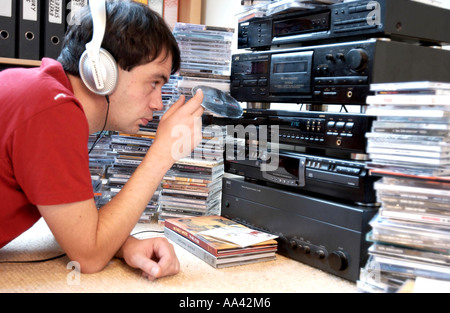 Giovane uomo nelle cuffie il caricamento di un cd in un sistema stereo con pile di album musicali tutti intorno a lui Foto Stock