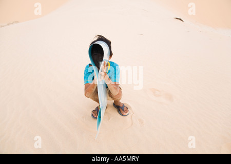 Mui Ne, Vietnam. Giovane ragazzo vietnamita scherzosamente holding slitta di sabbia a dune di sabbia bianca Foto Stock