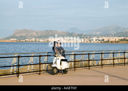 Coppia giovane baciando su una Vespa accanto al mare in prima serata, Palermo, Sicilia, Italia Foto Stock