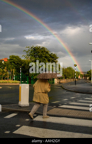 Donna che cammina attraverso le strisce pedonali con ombrellone e rainbow nel grigio cielo nuvoloso Foto Stock