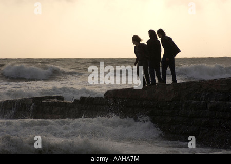 Silhouette di tre persone giovani uomini in piedi sul molo di pietra durante una tempesta schivare le onde ; rischi per la salute e la sicurezza Foto Stock