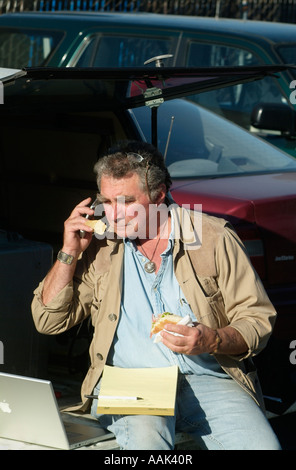 Fotografo colloqui sul suo telefono cellulare, mentre mangia il pranzo sul retro del suo carrello. Foto Stock