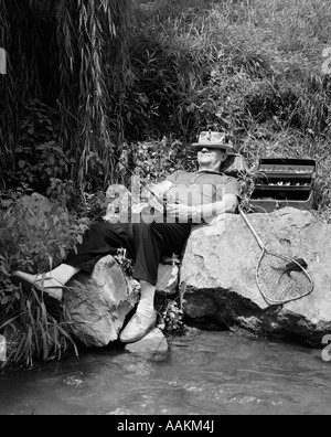 Anni Cinquanta LAZY FISHERMAN mentire indietro sulla roccia con HAT tirata sopra gli occhi la pesca in fiume Foto Stock