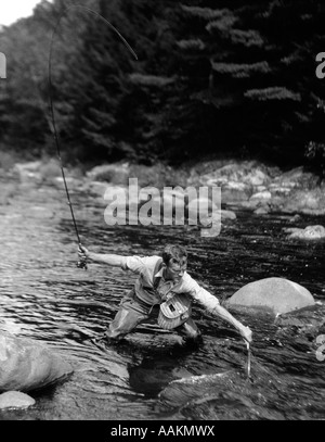 1920s uomo nel flusso indossando WADERS con pesce sulla linea cercando di catturare IN NET Foto Stock