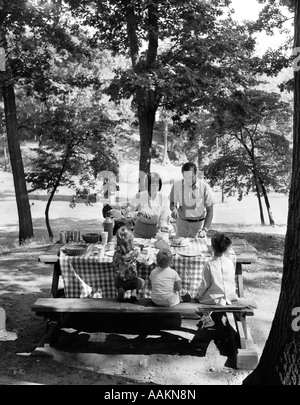 Anni sessanta anni settanta famiglia di cinque persone a tavola nel parco sotto alberi con picnic Foto Stock