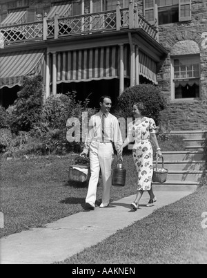 1930s giovane a piedi fuori di casa giù il marciapiede che porta cestini da picnic & caraffa thermos Foto Stock