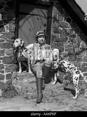 1930s sportiva alla moda donna seduta sulle fasi di costruzione in pietra con due cani dalmata Foto Stock