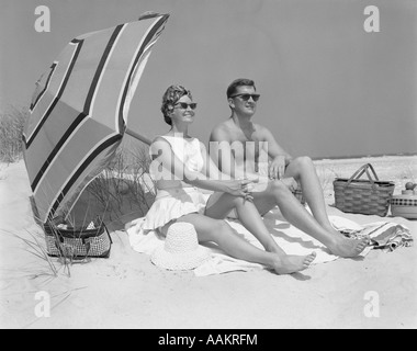 Anni sessanta paio di occhiali da sole seduto sulla spiaggia coperta con le gambe estese con ombrellone Foto Stock