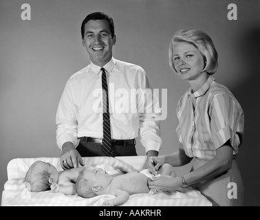 Anni sessanta sorridendo IL PADRE E LA MADRE CON TWIN neonati sul pannolino MODIFICA TABELLA guardando la fotocamera Foto Stock