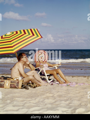 Anni Cinquanta anni sessanta la mezza età giovane seduto sulla spiaggia sotto ombrello uomo con mazze da golf sembra annoiato moglie felice Foto Stock