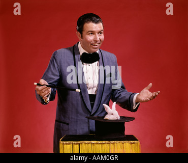 Anni Settanta uomo mago sventolando la bacchetta su top hat capovolta sul tavolo bianco appare di coniglio Foto Stock