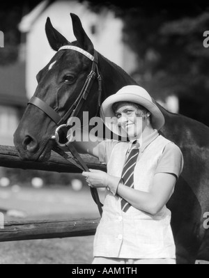 1930s donna in piedi accanto a cavallo azienda redini sorridente indossando di moda HAT STRIPED Tie collo guanti giubbotto di biancheria GUARDANDO Foto Stock