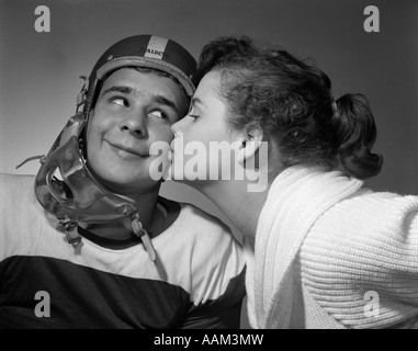 Anni Cinquanta TEEN GIRL coda di cavallo baciare Ragazzo indossa casco da football americano sulla guancia Foto Stock