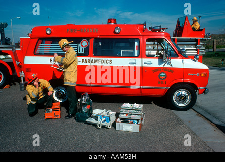 Anni Settanta anni ottanta due uomini del team paramedico ISPEZIONE DI CONTROLLO APPARECCHIATURE IN AMBULANZA VAN VIGILI DEL FUOCO che indossa caschi GIACCHE Foto Stock