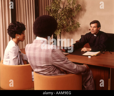 1970 anni settanta sacerdote ministro clero uomo intervistando CONSULTING AFRICAN-AMERICAN GIOVANE UOMO DONNA DESK OFFICE RELIGIONE Foto Stock