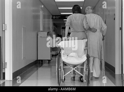 Anni ottanta VISTA POSTERIORE DELLA DONNA CON IL BRACCIO intorno all uomo anziano utilizzando WALKER SEDIA A ROTELLE dietro di loro Foto Stock