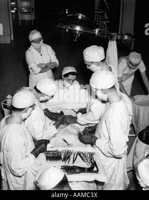 1930s équipe chirurgica indossando abiti bianchi CAPPELLI E MASCHERE IN OSPEDALE IN SALA OPERATORIA eseguendo una procedura medica Foto Stock