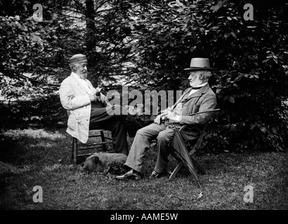 Giro del secolo due uomini seduti fuori in sedie tra alberi di fumare sigari cane parlante recante AI LORO PIEDI Foto Stock