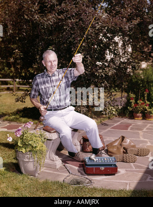 Anni sessanta vecchio uomo seduto in cortile con la canna da pesca E INGRANAGGIO TACKLE BOX CANTRA STIVALI NET PENSIONATO HOBBY Foto Stock